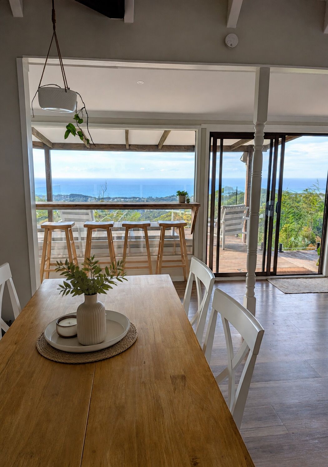 Un comedor con una mesa de madera, sillas blancas y vista al océano a través de grandes puertas de cristal.