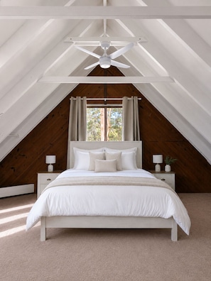 A bedroom with a sloped ceiling, a bed with white bedding, two bedside tables with lamps, and a window with curtains.