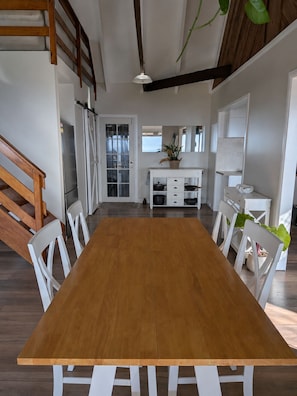 A dining area with a wooden table and white chairs, a kitchen in the background, and a staircase with wooden railings.