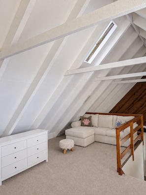 A loft bedroom with a sloped ceiling, a white dresser, a beige sofa, a small ottoman, and a wooden ladder.