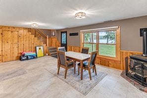 A spacious room with a wooden staircase, a dining table with chairs, a fireplace, and a large window offering a view of the outdoors.