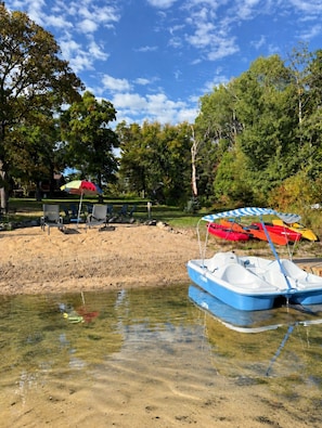 A clear lake with a sandy shore, a rowboat, and beach chairs under umbrellas.