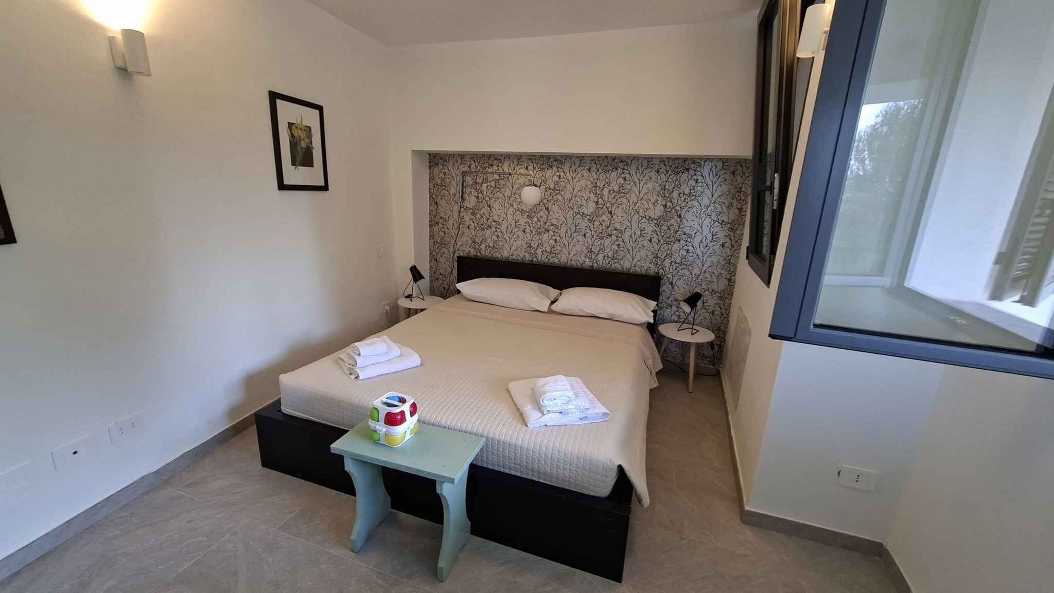 Un lit bien fait, avec des oreillers et une serviette pliée, une petite table sur laquelle se trouvent une tasse et un jouet, et un tableau accroché au mur.
