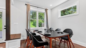 Ein Esszimmer mit einem rechteckigen Tisch, gedeckt für vier Personen, umgeben von schwarzen Stühlen, einem Fenster mit Vorhängen und Blick auf Grünfläche.