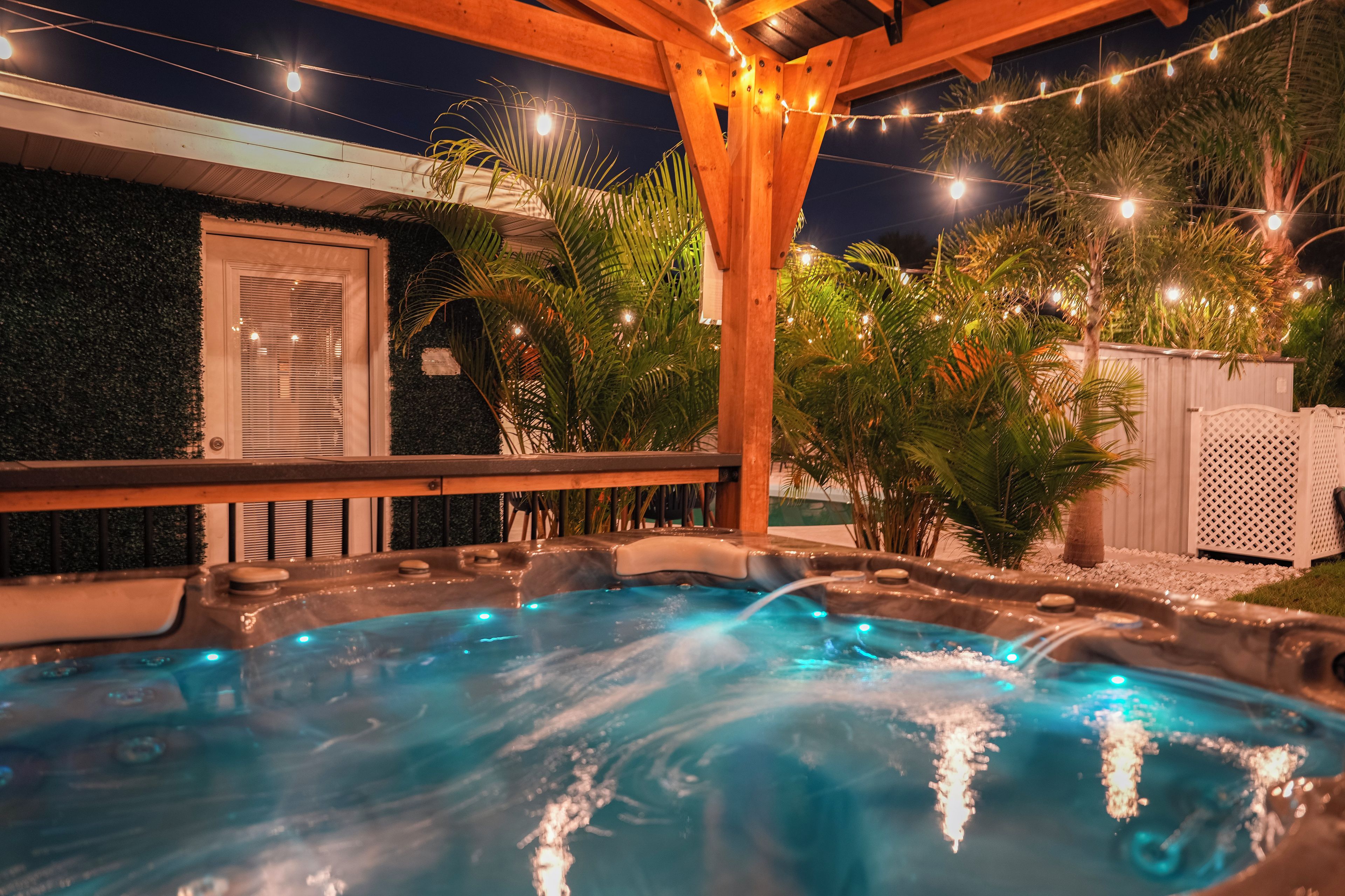 Ein Whirlpool mit Holzterrasse und Lichterkette, umgeben von Grünpflanzen und einem weißen Zaun.
