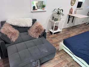 Una sala de estar moderna con un sofá gris, almohadones mullidos, una pequeña mesita auxiliar blanca y una cama azul.