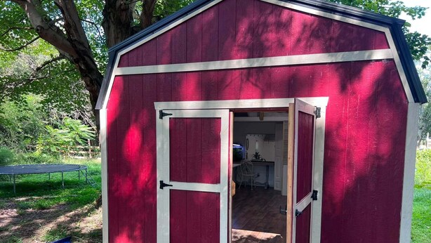 Una estructura de madera roja con una puerta y ventanas, rodeada de árboles y grava.