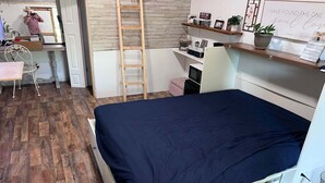 Un dormitorio con cama, escritorio, silla, escalera y estante con diversos objetos.