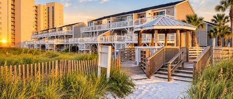 Un hotel frente a la playa con varios balcones, un espacio exterior cubierto y un camino de arena que lleva a la entrada.