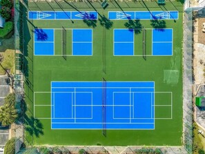 Vista aérea de un complejo de canchas de tenis con múltiples canchas, rodeado de vegetación y una cerca.