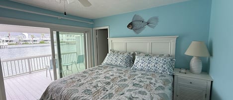 A bedroom with a bed, a nightstand, a lamp, a ceiling fan, and a view of a water body through a sliding door.