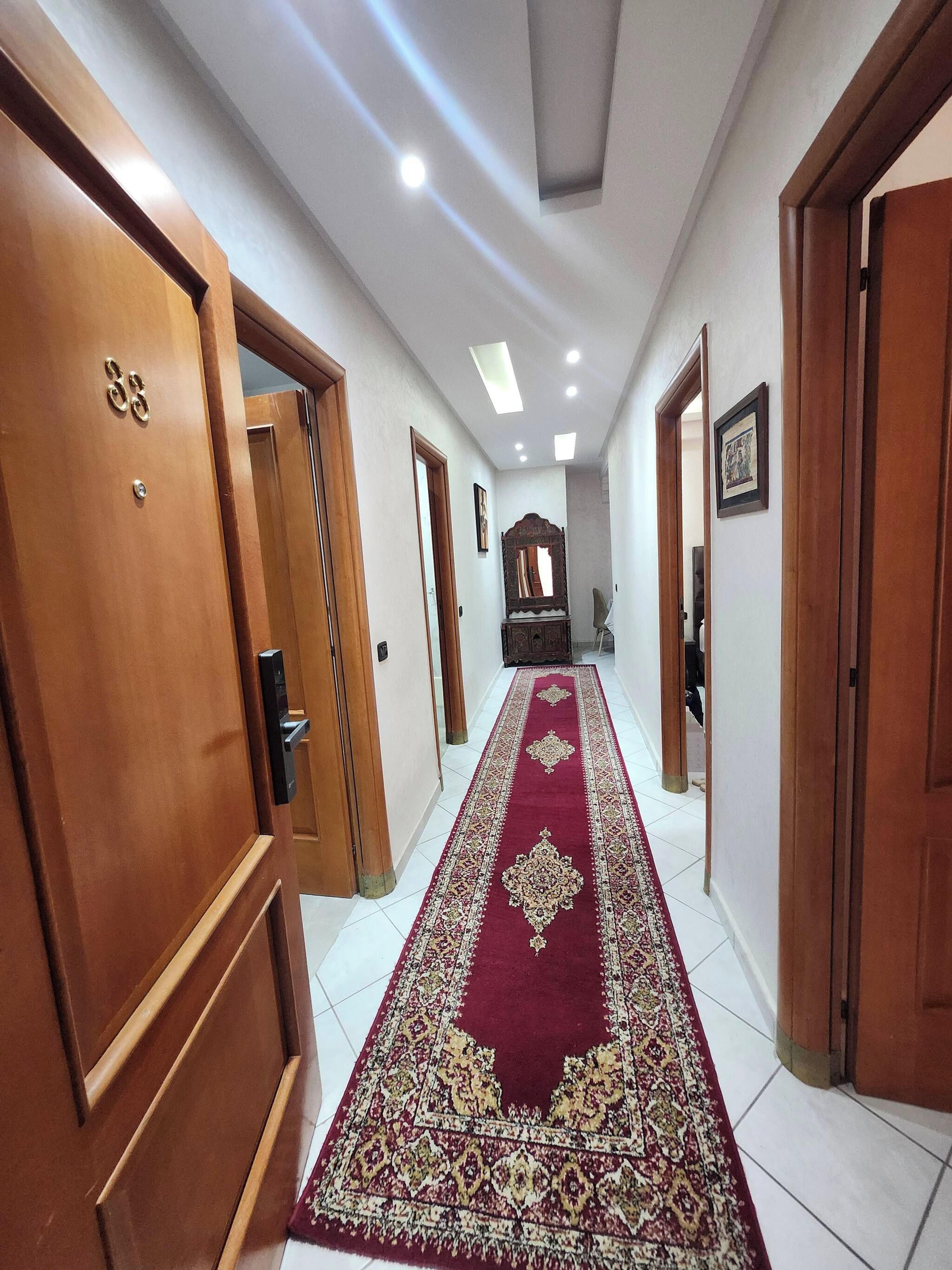 Un pasillo con puertas de madera, alfombra roja estampada e iluminación empotrada en el techo.