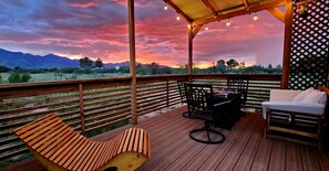 Un terrazzo in legno con una poltrona da relax, un tavolino e un divano, con vista su un paesaggio suggestivo tra montagne e tramonto.