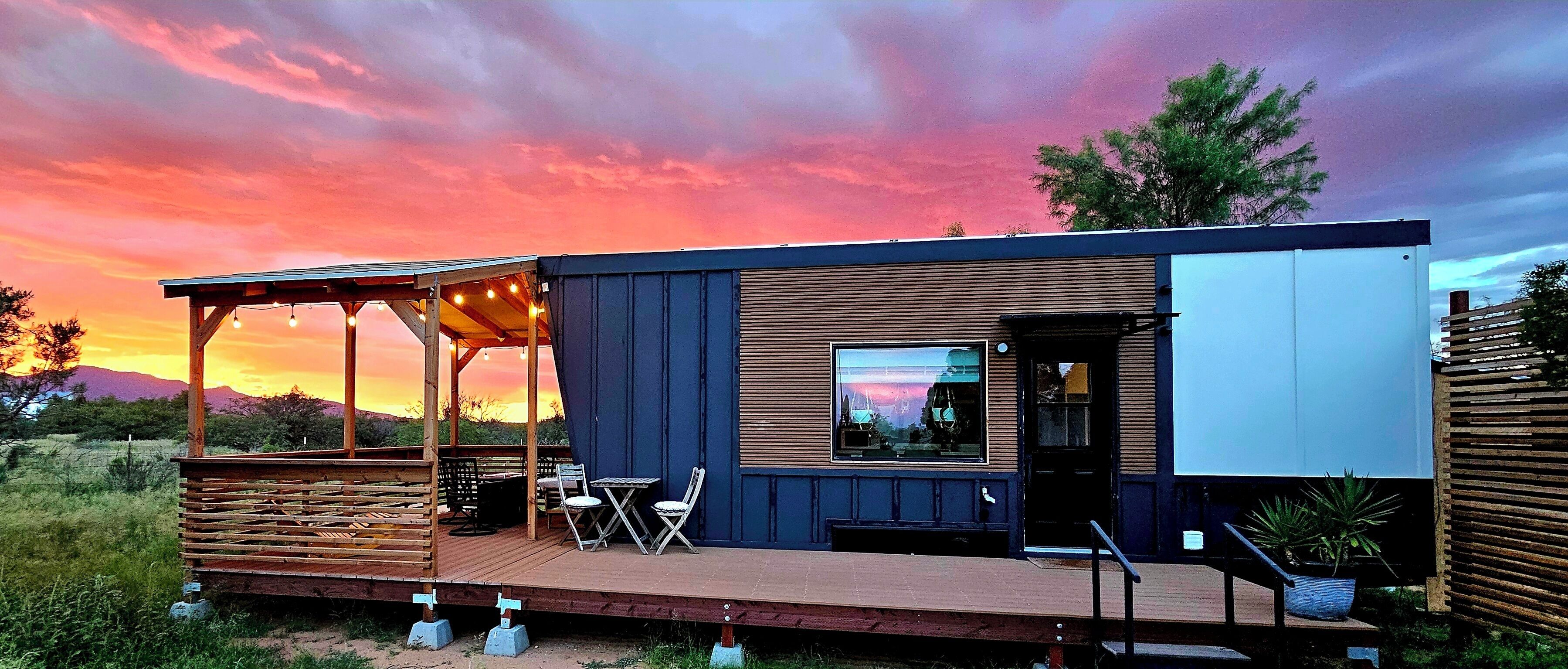 A modern, compact cabin with a deck, outdoor seating, and a large window.