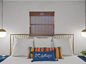 Ein Bett mit weißer Bettwäsche, ein blaues Kopfkissen mit der Aufschrift 'ocean' und ein Fenster mit Jalousien.