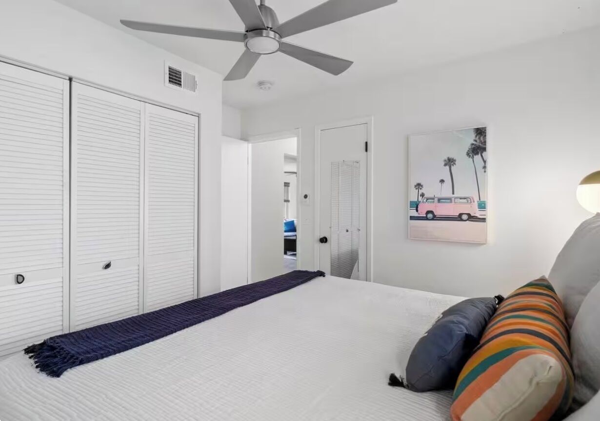 Una camera da letto con un letto, una ventola a soffitto, una porta e un quadro appeso al muro.