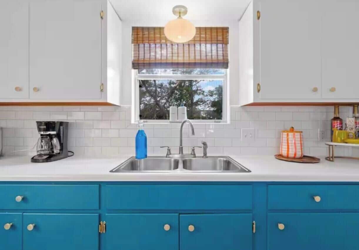 Una cucina con mobili bianchi, un piano di lavoro blu, una finestra con persiane, una macchina da caffè e un lavello.