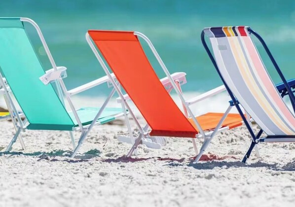 Three beach chairs with metal frames and fabric seats, arranged on a sandy beach with a clear view of the ocean in the background.