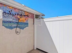 A mosaic wall with a showerhead, a white fence, and a clear blue sky.