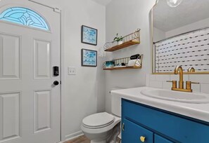 Un baño con inodoro blanco, tocador azul, lavabo dorado y estantes con adornos.