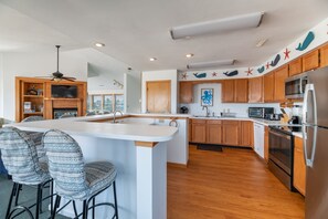 A modern kitchen with wooden cabinets, a large island with bar stools, and a view of the outdoors through large windows. - Sandpiper - Whalehead, Semi-Oceanfront, Pool, Hot Tub, Pets OK, YR Flex Stays (Corolla)