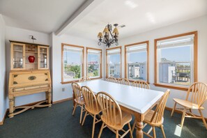 A dining room with a large table and chairs, a wooden cabinet with glass doors, and multiple windows offering views of the outside. - Sandpiper - Whalehead, Semi-Oceanfront, Pool, Hot Tub, Pets OK, YR Flex Stays (Corolla)