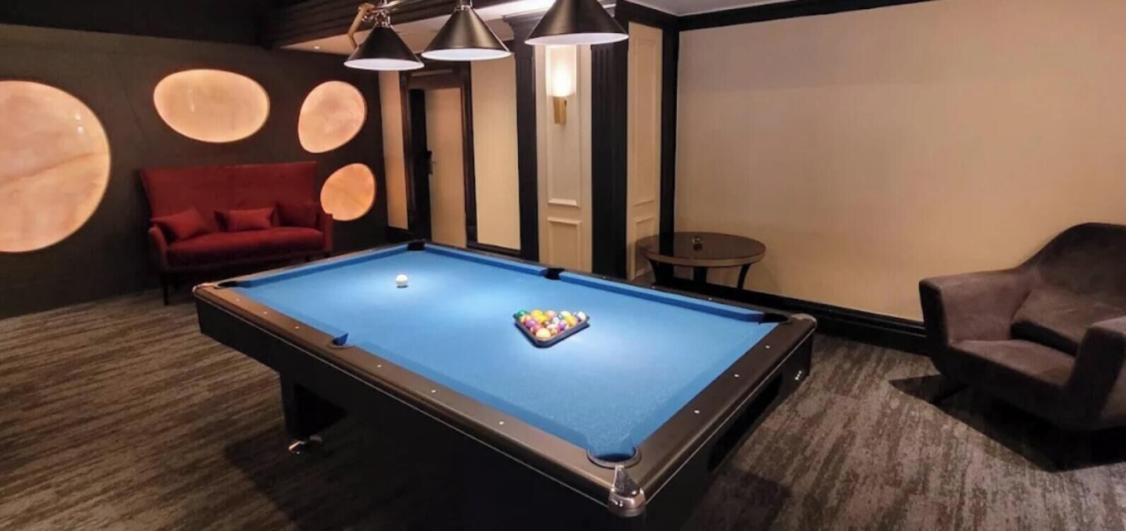 Ein Billardtisch mit einem roten Sofa und einem kleinen runden Tisch in einem Raum mit dunklen Wänden und Deckenleuchten.