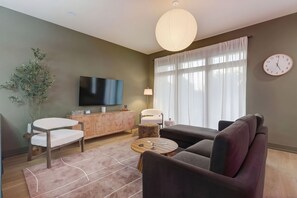 Ein modernes Wohnzimmer mit einem Flachbildfernseher, einer Couch, einem Sessel, einem kleinen runden Tisch und einem großen Fenster mit Vorhängen.
