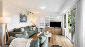 A modern living room with a sofa, a coffee table, a flat-screen TV, and a large window with shutters. - Ocean Shores 10 Peregian Beach (Peregian Beach)