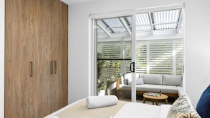 A modern bedroom with a wooden wardrobe, a sofa, a small round table, and a sliding glass door leading to a balcony with a view of greenery. - Ocean Shores 10 Peregian Beach (Peregian Beach)