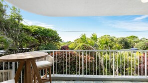 A balcony with a wooden table and chairs, surrounded by a white railing, overlooking a lush garden with various trees and plants. - Ocean Shores 10 Peregian Beach (Peregian Beach)