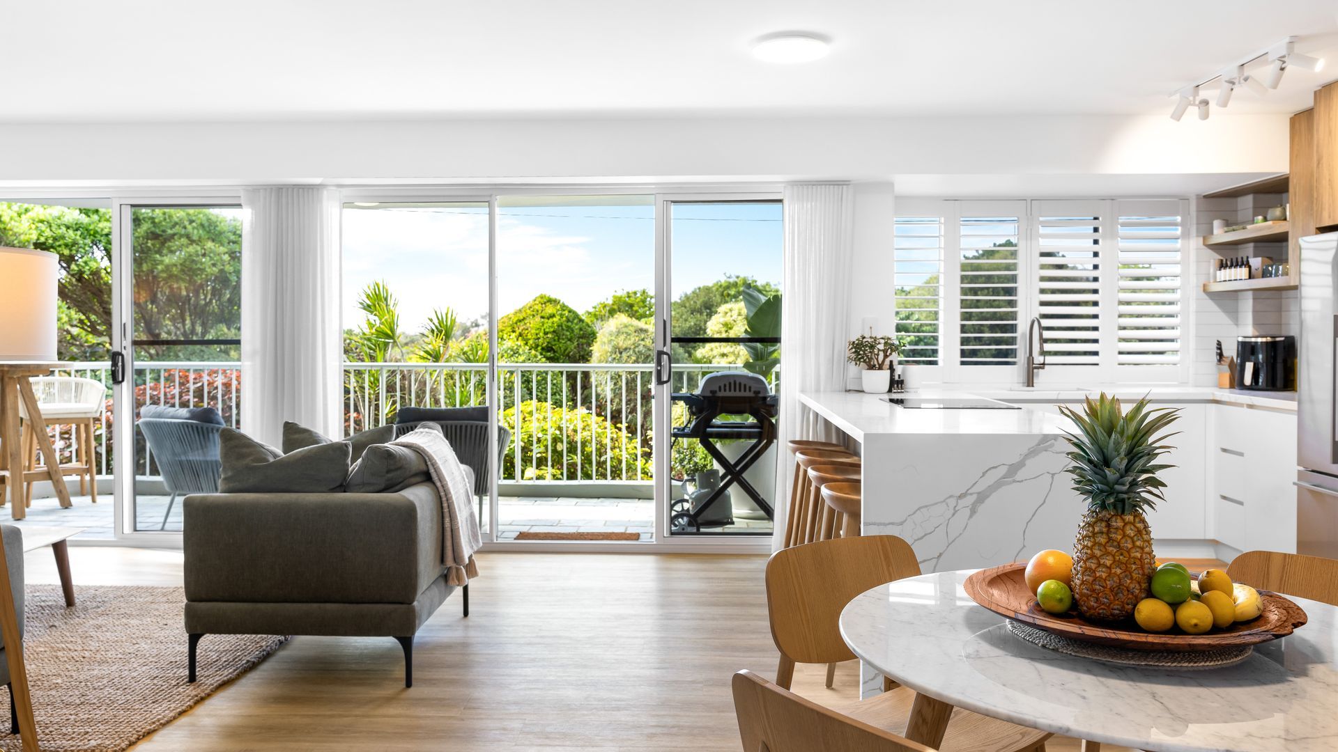 A modern kitchen with a large island, a dining area with a round table, and a balcony with a view of greenery.