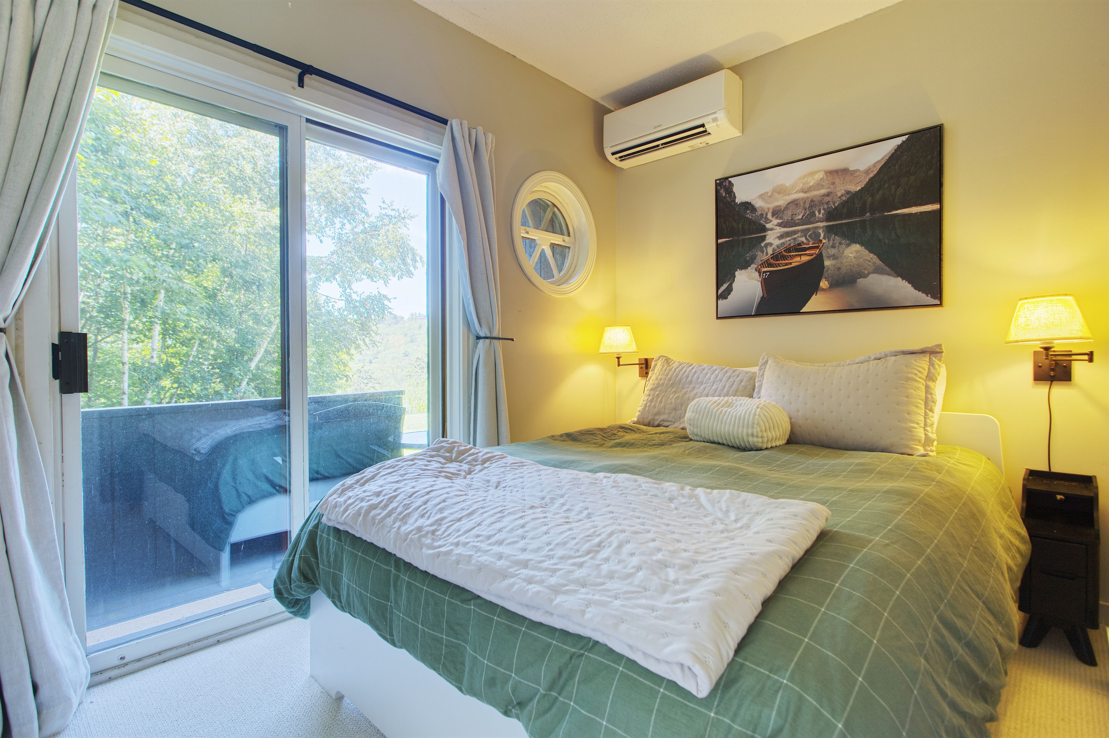 A bedroom with a bed, two bedside lamps, a window with curtains, a wall-mounted air conditioner, and a painting of a boat on water.