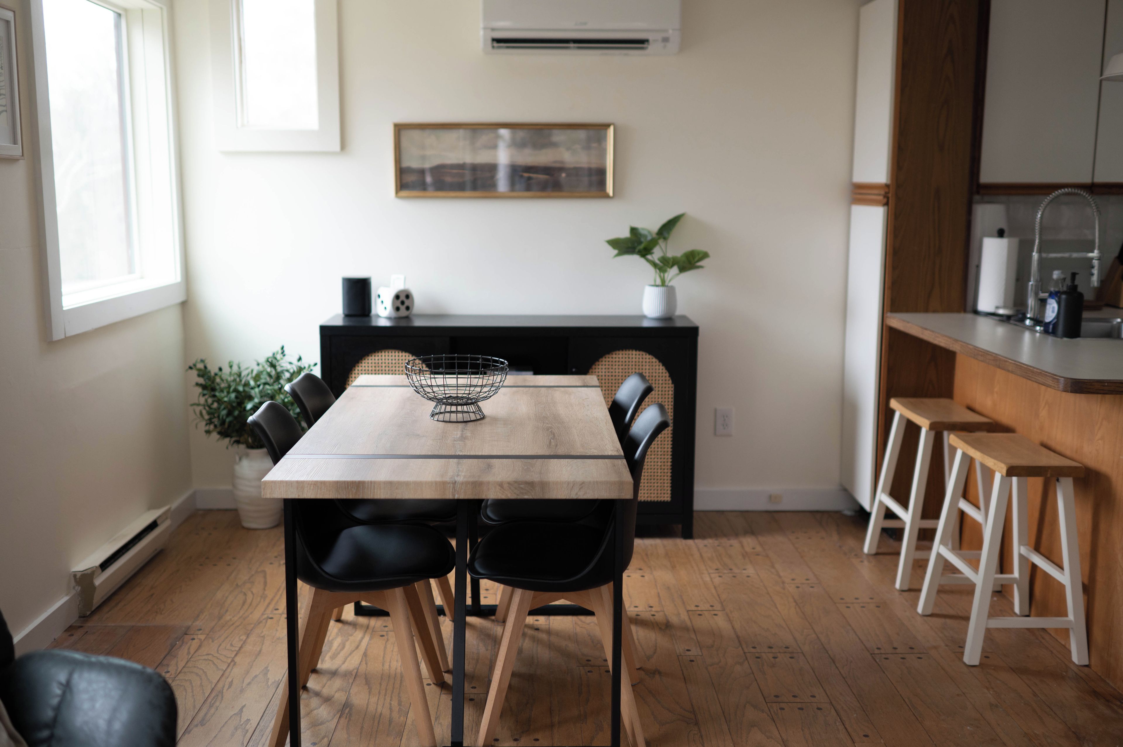 A dining area with a wooden table, black chairs, a black console table with a plant, and a kitchen area with stools.