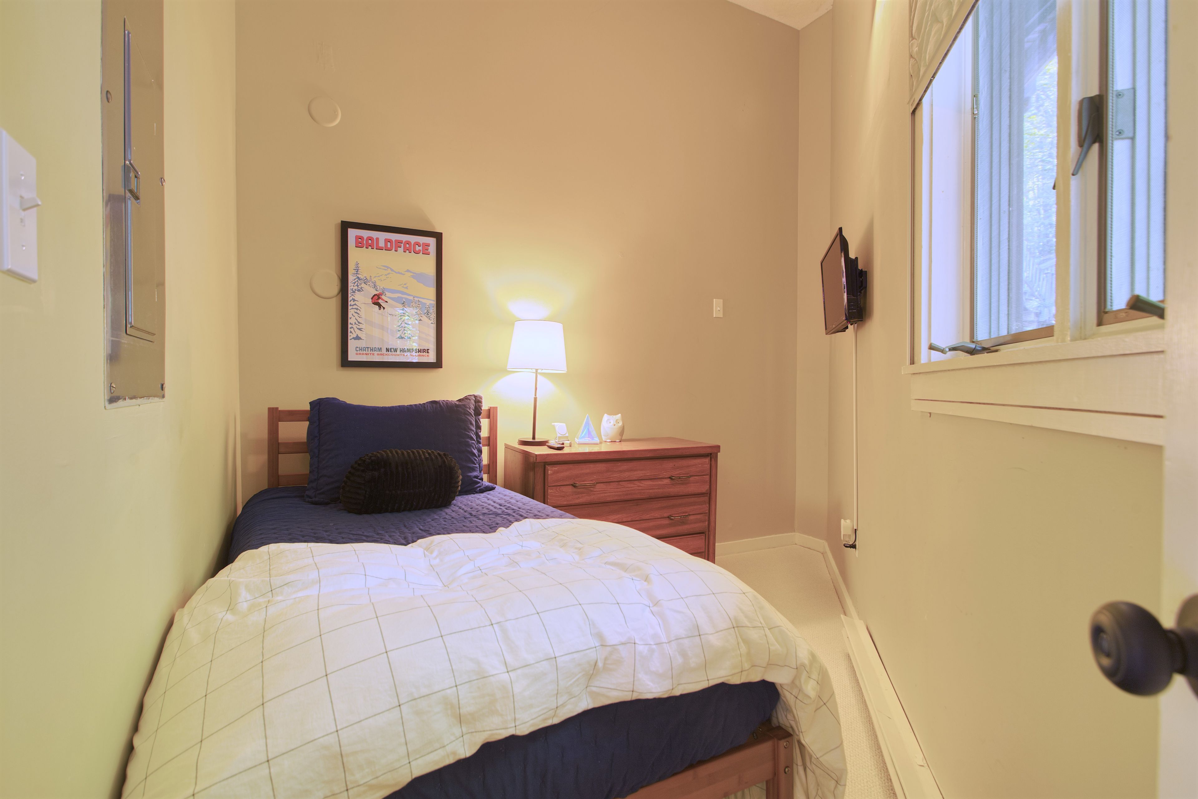 Une chambre à coucher avec un lit, une table de chevet, une lampe, une affiche au mur et une fenêtre.