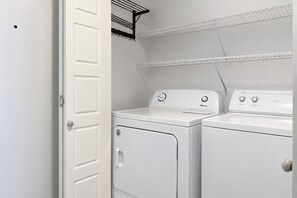 A laundry room with two white front-loading washers and dryers, a metal drying rack, and a white door. - Gulf Front | Huge Balcony | Direct Beach Access | BEACHBALL PROPERTIES (Orange Beach)