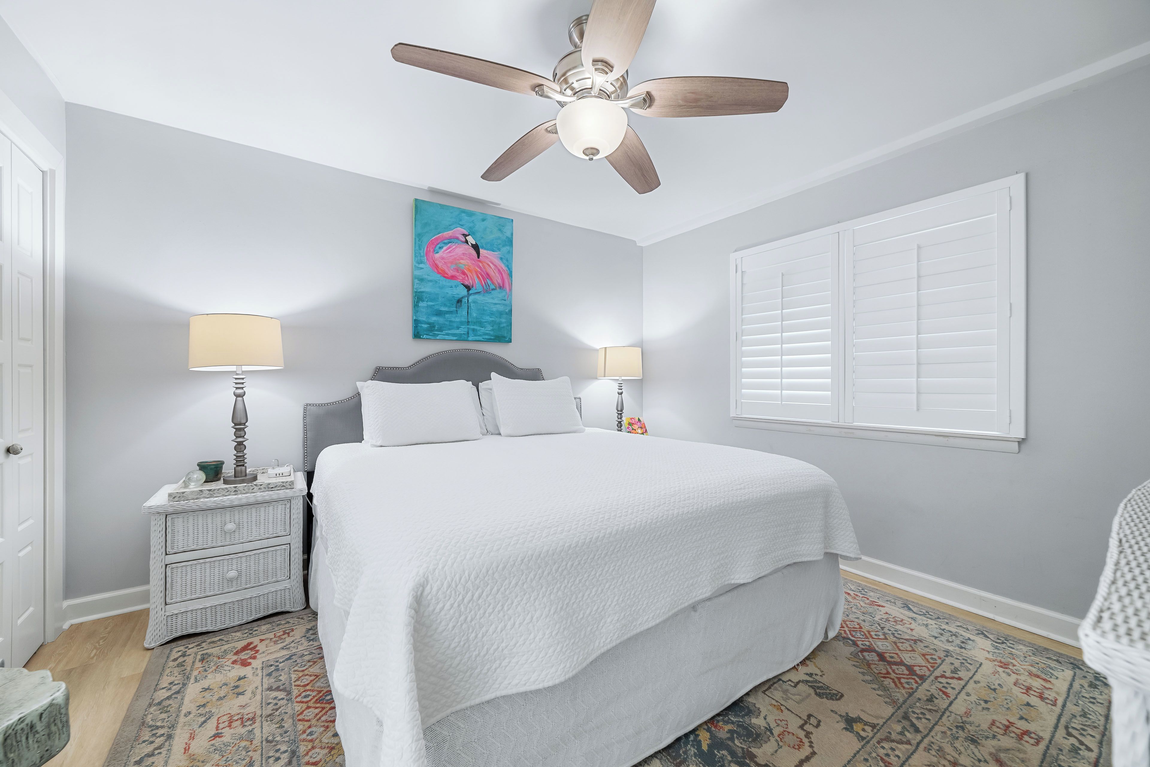 A bedroom with a bed, two bedside lamps, a ceiling fan, a painting, and a window with blinds.