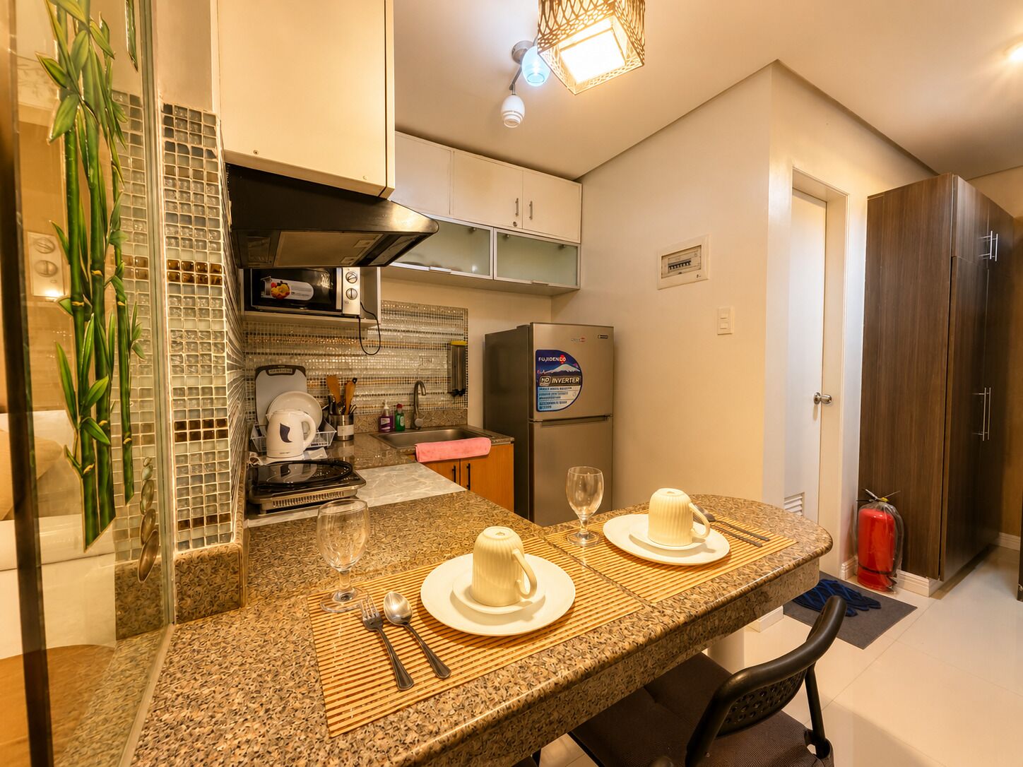 A compact kitchen with a microwave, stove, and refrigerator. There is a dining table set with plates, cutlery, and glasses.