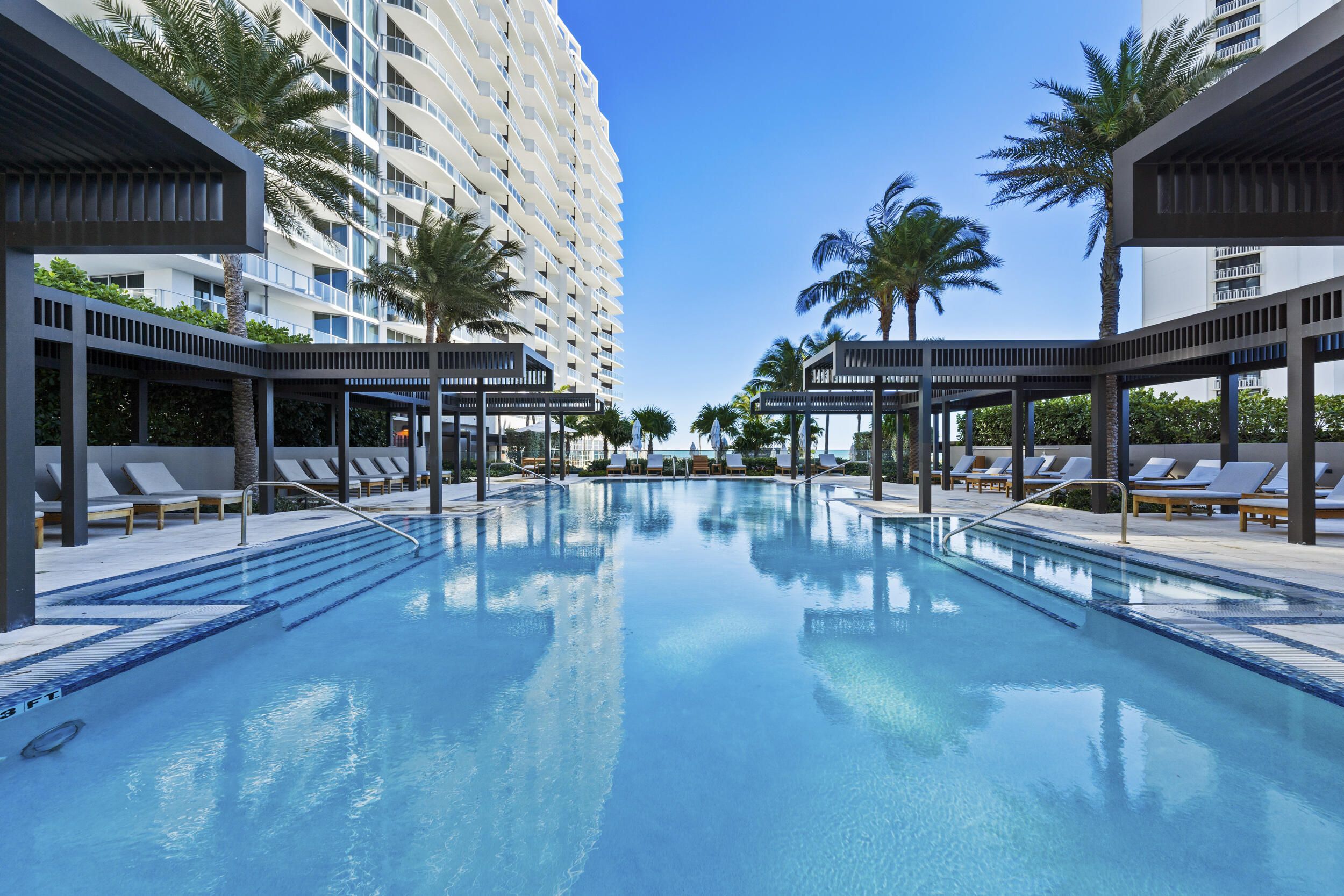 A swimming pool with lounge chairs and palm trees, surrounded by modern buildings.