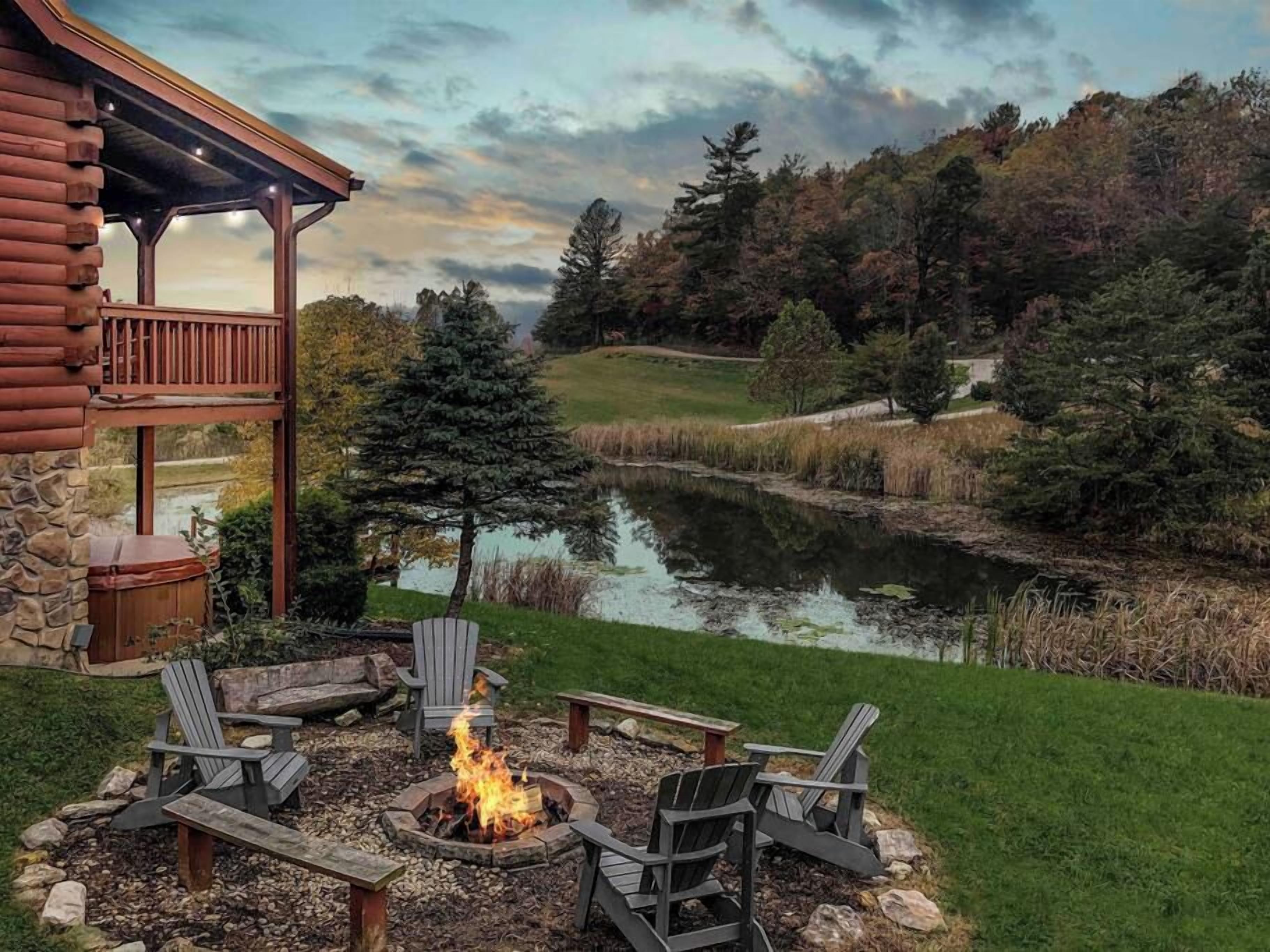 A cabin with a fire pit and Adirondack chairs, a stone fireplace, and a view of a pond and forest.