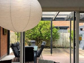 A covered patio area with a table and chairs, a hanging light fixture, and a view of a backyard with a fence and trees. - 3BR Belmont House | Walk to High St Shops (Belmont)