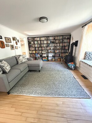 Ein Wohnzimmer mit grauem Ecksofa, einem großen Bücherregal voller Bücher, einem Kamin und einem Fenster mit Vorhängen.