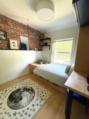 Ein Schlafzimmer mit Backsteinwand, einem Fenster mit Blick, einem Bett, einem Schreibtisch und einem runden Teppich mit Sternen.