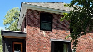 Une maison à deux étages, construite en briques, avec une porte d’entrée, une boîte aux lettres et un petit jardin devant, bordé d’une haie.