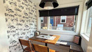 Un coin repas avec une table en bois, des chaises, un banc et une vue sur un bâtiment en briques, visible à travers une fenêtre.