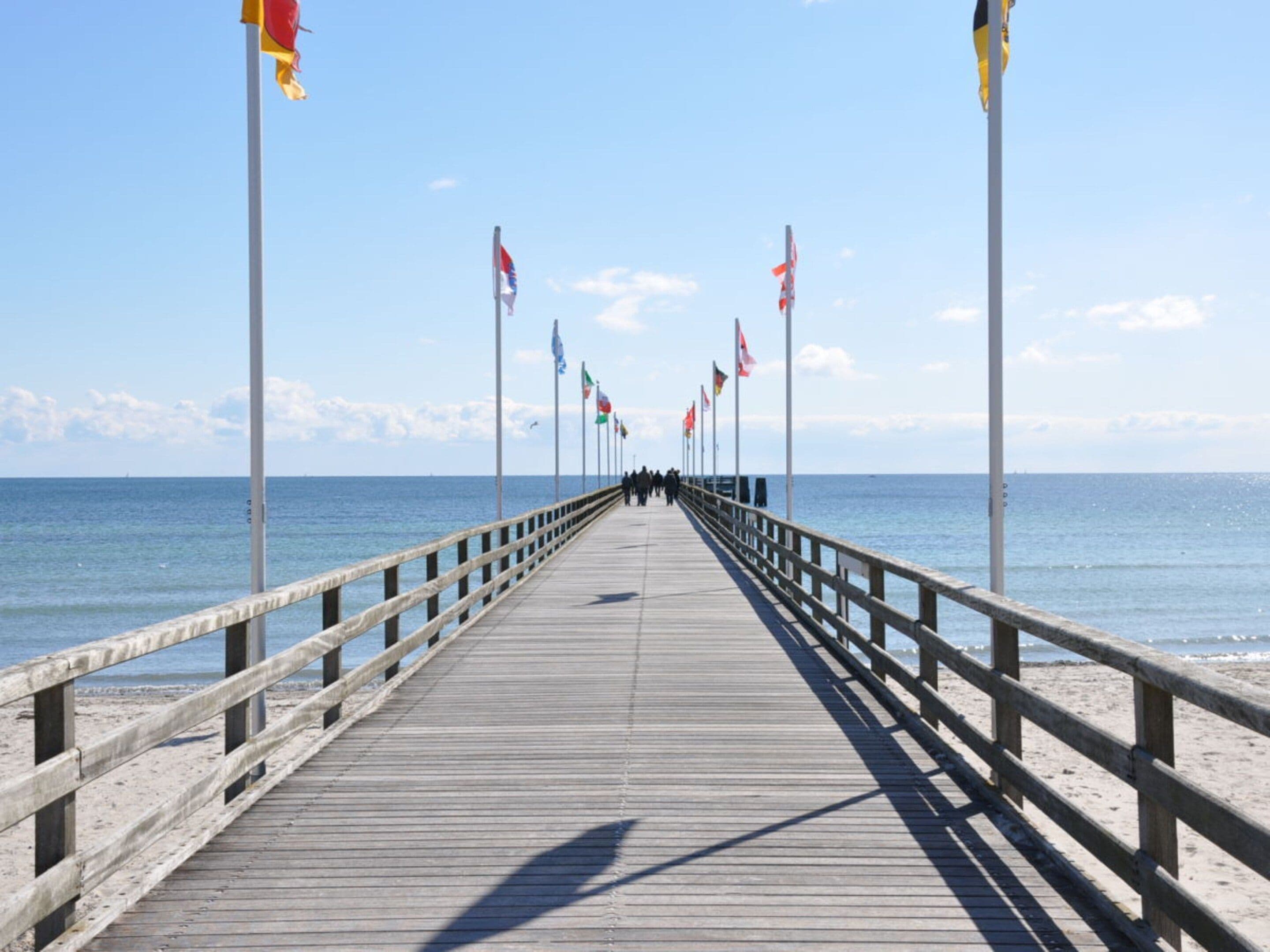 Un muelle de madera se adentra en el mar con banderas en mástiles, conduciendo a una playa de arena.