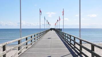 Un muelle de madera se adentra en el mar con banderas en mástiles, conduciendo a una playa de arena.