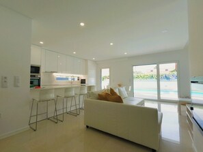 A modern kitchen with white cabinets, a bar area, and a large window overlooking a pool. - Aroeira Nest | 3-Bedroom Villa with Pool and BBQ (Almada)