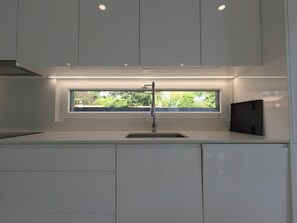 A modern kitchen with a window above the sink, recessed lighting, and white cabinetry. - Aroeira Nest | 3-Bedroom Villa with Pool and BBQ (Almada)