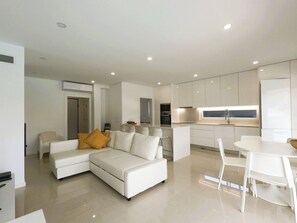 A modern living room with a white sofa, a dining table, and a kitchen area with white cabinets and a built-in oven. - Aroeira Nest | 3-Bedroom Villa with Pool and BBQ (Almada)